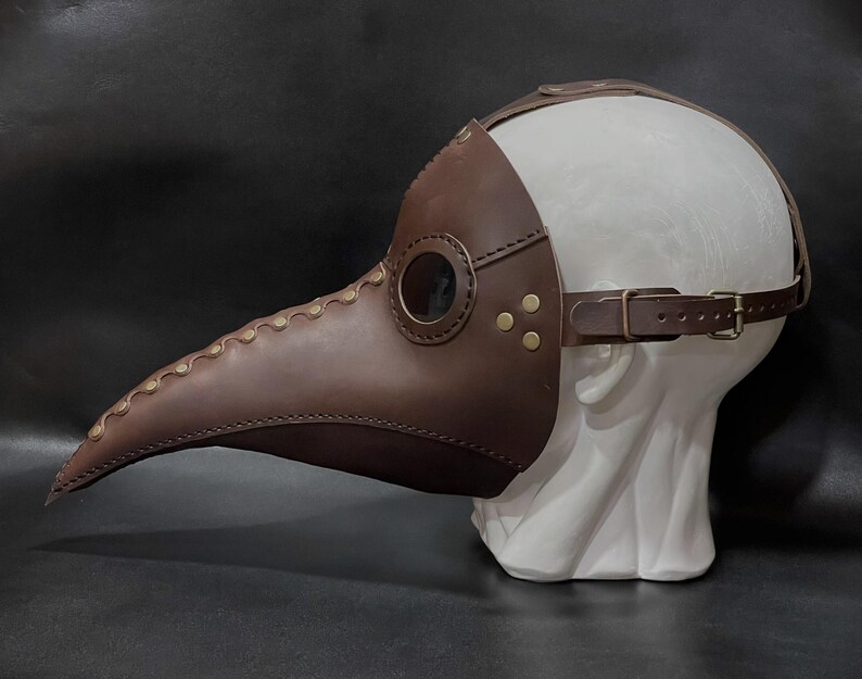 May include: A brown leather plague doctor mask with a long beak and eye holes. The mask is on a white mannequin head.