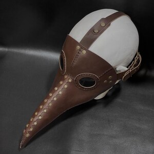 May include: A brown leather plague doctor mask with a long beak and eye holes. The mask has brass rivets and a leather strap for wearing.