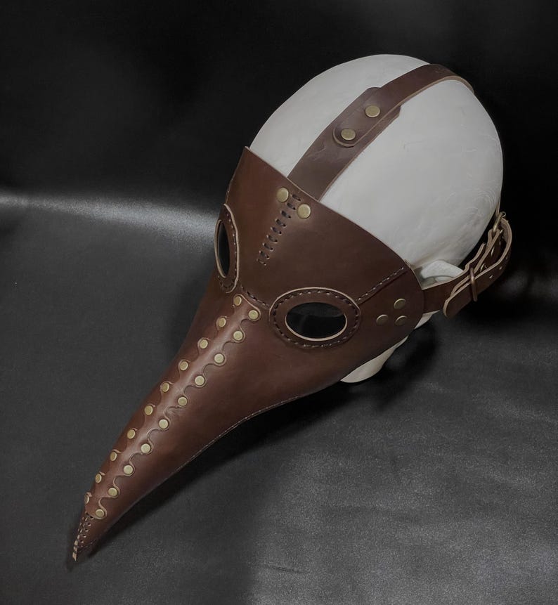 May include: A brown leather plague doctor mask with a long beak and eye holes. The mask has brass rivets and a leather strap for wearing.