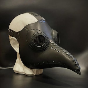 Peut inclure: Un masque de médecin de la peste en cuir noir avec un long bec incurvé et des ouvertures oculaires circulaires. Le masque est orné de rivets et d'une sangle. Il est présenté sur une tête de mannequin blanche sur fond sombre.