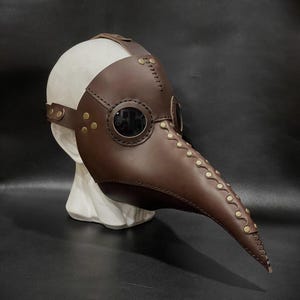 May include: A brown leather plague doctor mask with a long beak and two round eyeholes. The mask has gold-colored rivets and stitching.