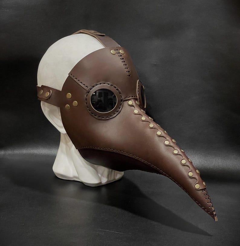 May include: A brown leather plague doctor mask with a long beak and two round eyeholes. The mask has gold-colored rivets and stitching.
