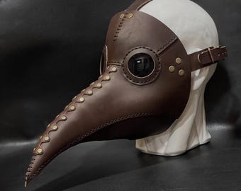 Könnte beinhalten: Eine braune Ledermaske eines Pestarztes mit einem langen Schnabel und Augenlöchern. Die Maske ist mit Messingnieten und Nähten verziert.