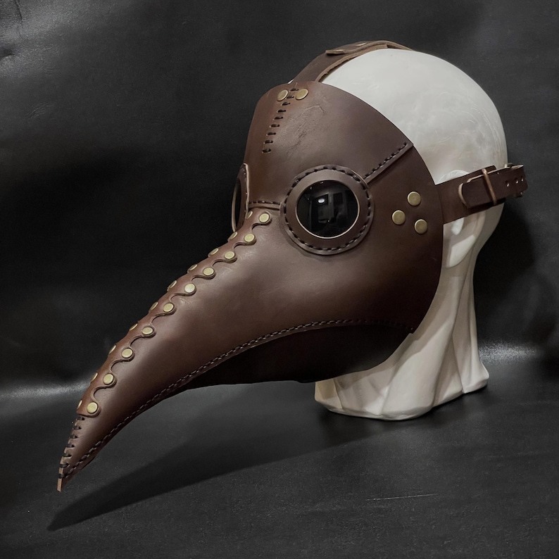 May include: A brown leather plague doctor mask with a long beak and eye holes. The mask is decorated with brass rivets and stitching.