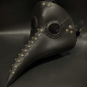 May include: A black leather plague doctor mask with a long beak and two round eyeholes. The mask is decorated with gold-colored rivets and stitching.