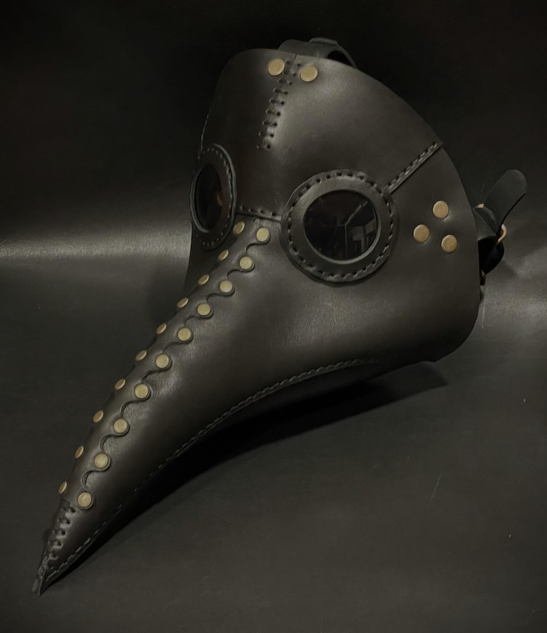 May include: A black leather plague doctor mask with a long beak and two round eyeholes. The mask is decorated with gold-colored rivets and stitching.