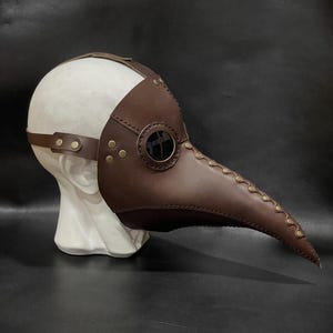 May include: A brown leather plague doctor mask with a long beak and a round glass eye piece. The mask is on a white mannequin head.