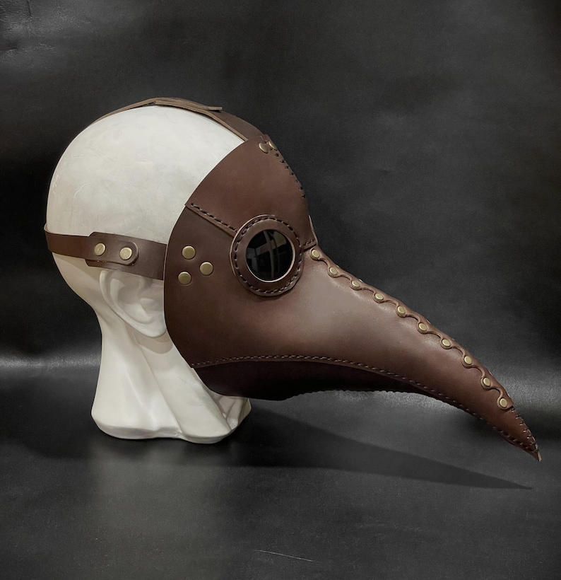 May include: A brown leather plague doctor mask with a long beak and a round glass eye piece. The mask is on a white mannequin head.