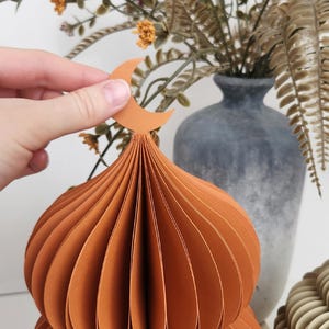 May include: An orange paper mosque dome with a crescent moon on top. The dome is made of layered paper and has a textured appearance. A hand holds the moon. A stone vase and dried flowers are in the background.