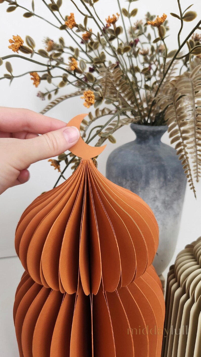 May include: An orange paper mosque dome with a crescent moon on top. The dome is made of layered paper and has a textured appearance. A hand holds the moon. A stone vase and dried flowers are in the background.