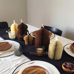 May include: Table setting featuring decorative paper 3D mosques in earth tones of brown and cream colors, shaped like domes, with crescent moon accents. The table is set with wooden plates, gold cutlery, and candles, creating an elegant and festive atmosphere.