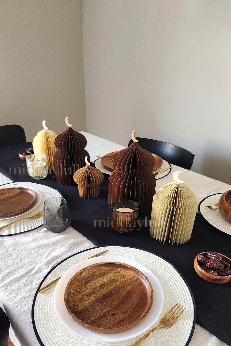 May include: Table setting featuring decorative paper 3D mosques in earth tones of brown and cream colors, shaped like domes, with crescent moon accents. The table is set with wooden plates, gold cutlery, and candles, creating an elegant and festive atmosphere.