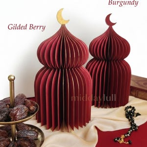 May include: Two burgundy paper mosques with gold crescent moon toppers. The mosques have a layered design and are displayed on a red surface with a gold serving tray of dates and black prayer beads with Quran in the back.