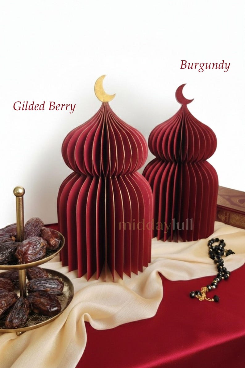 May include: Two burgundy paper mosques with gold crescent moon toppers. The mosques have a layered design and are displayed on a red surface with a gold serving tray of dates and black prayer beads with Quran in the back.