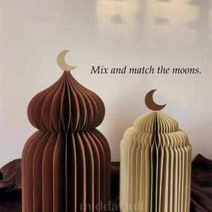 May include: Two paper dome-shaped decorations, one brown and one cream, each topped with a crescent moon. The brown dome has a light beige moon, and the cream dome has a dark brown moon. Text reads "Mix and match the moons."