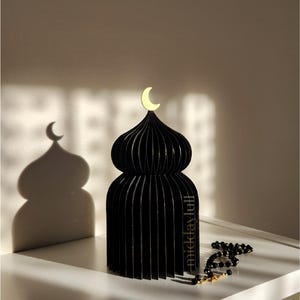 May include: Black decorative 3D mosque with gilded edges and a gold crescent moon on top. The mosque is made of layered material, casting a beautiful shadow. A string of black prayer beads with gold accents rests nearby. The text "Starry Night" is visible.