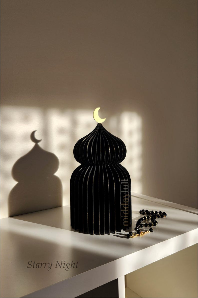 May include: Black decorative 3D mosque with gilded edges and a gold crescent moon on top. The mosque is made of layered material, casting a beautiful shadow. A string of black prayer beads with gold accents rests nearby. The text "Starry Night" is visible.