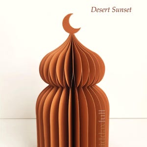 May include: A brown paper sculpture of a domed structure, possibly a mosque, with a crescent moon on top. The text "Desert Sunset" is visible. The design is made of layered, folded paper, creating a 3D effect.