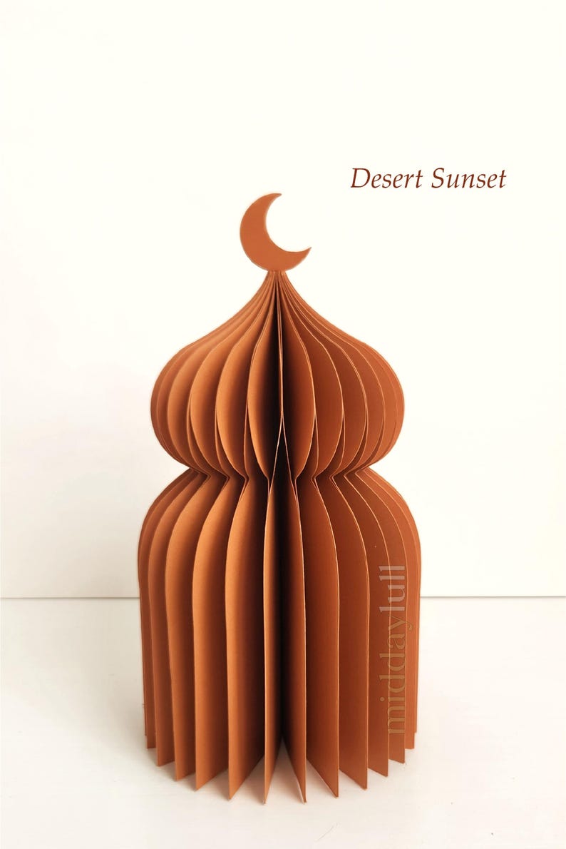 May include: A brown paper sculpture of a domed structure, possibly a mosque, with a crescent moon on top. The text "Desert Sunset" is visible. The design is made of layered, folded paper, creating a 3D effect.