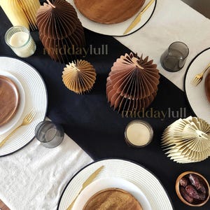 May include: A festive table setting with white plates, wooden chargers, gold cutlery, and decorative 3D paper mosques in shades of brown and cream. The table is set with candles and small glasses, creating an elegant dining arrangement.