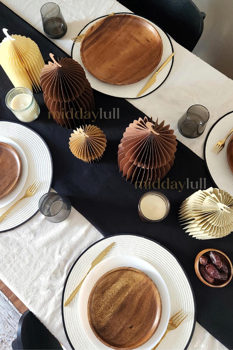 May include: A festive table setting with white plates, wooden chargers, gold cutlery, and decorative 3D paper mosques in shades of brown and cream. The table is set with candles and small glasses, creating an elegant dining arrangement.
