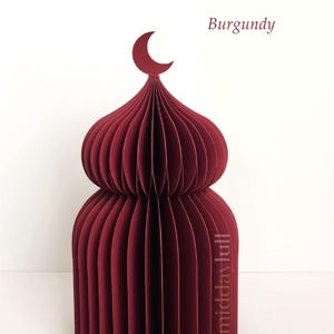 May include: A burgundy paper model of a mosque, featuring a crescent moon finial. The layered, curved paper construction forms a detailed architectural design. The word "Burgundy" is written in the upper right corner.