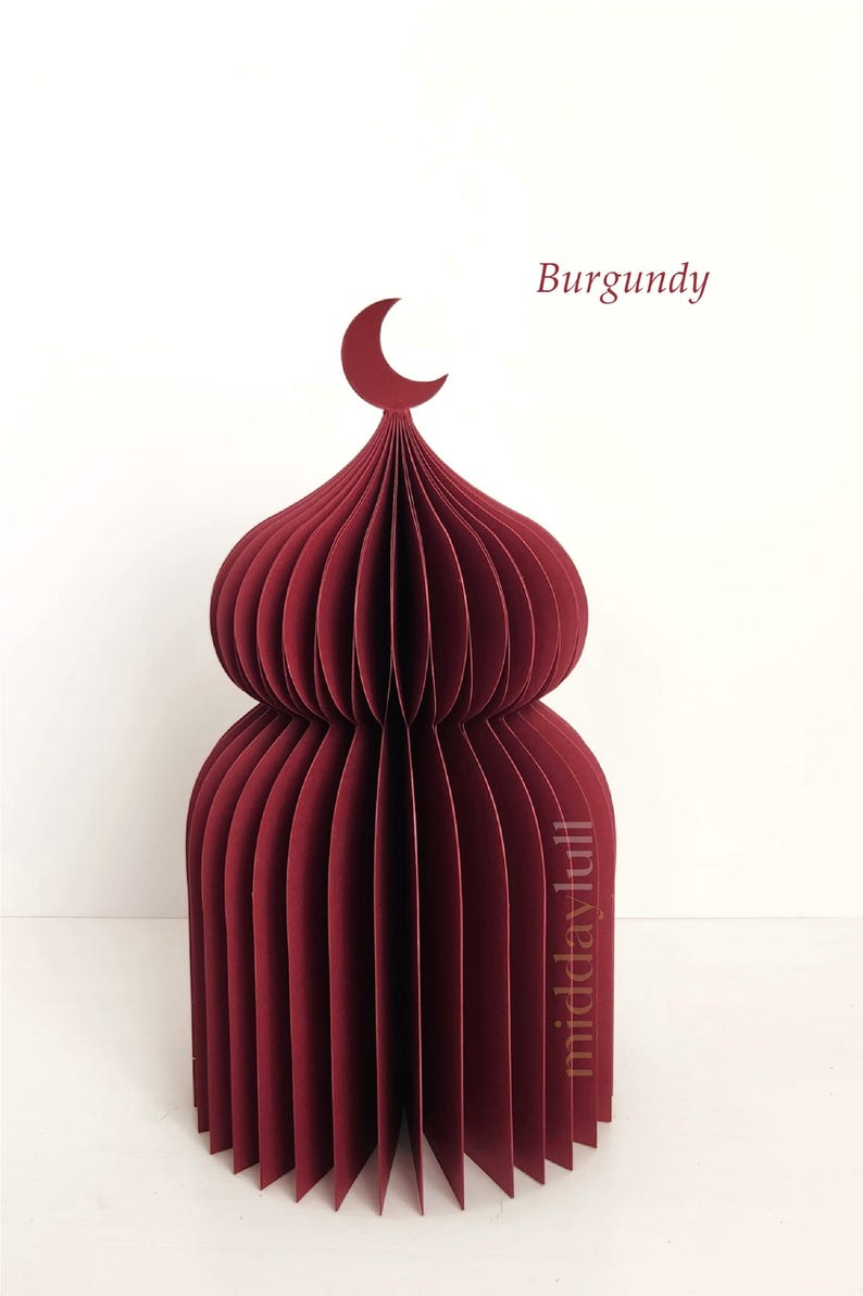 May include: A burgundy paper model of a mosque, featuring a crescent moon finial. The layered, curved paper construction forms a detailed architectural design. The word "Burgundy" is written in the upper right corner.
