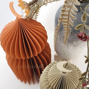 May include: Two 3D paper mosques in shades of orange and beige, with a crescent moon design. The mosques are made of folded paper and have a layered, textured appearance. The background includes a vase with dried flowers.
