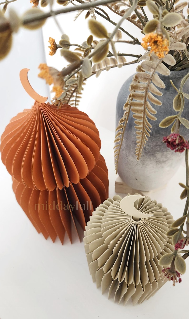 May include: Two 3D paper mosques in shades of orange and beige, with a crescent moon design. The mosques are made of folded paper and have a layered, textured appearance. The background includes a vase with dried flowers.