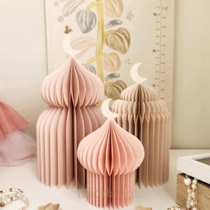 May include: Three paper mosque decorations in dusty rose and beige, topped with crescent moon cutouts. The text "Dusty Rose Bundle" is visible. Wooden star accents and a beaded bracelet are also present.