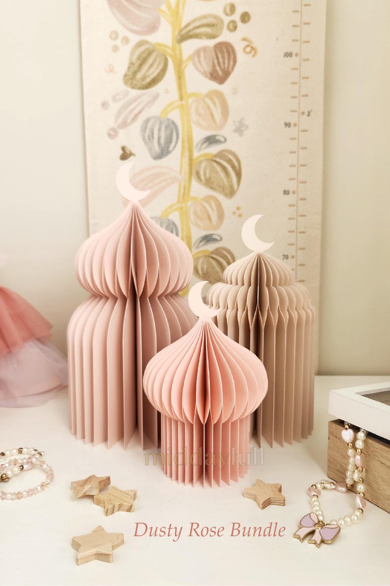 May include: Three paper mosque decorations in dusty rose and beige, topped with crescent moon cutouts. The text "Dusty Rose Bundle" is visible. Wooden star accents and a beaded bracelet are also present.