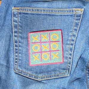 Könnte beinhalten: Nahaufnahme einer blauen Jeans-Tasche mit einem farbenfrohen, gestickten Tic-Tac-Toe-Spiel. Das Spielbrett ist hellblau mit einem rosa Rand, und die X's und O's sind gelb. Die Tasche hat gelbe Nähte.