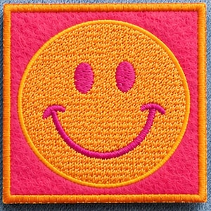 Könnte beinhalten: Ein quadratischer Stickaufnäher mit einem leuchtend orangefarbenen, kreisförmigen Smiley-Gesicht mit pinkfarbenen Augen und einem geschwungenen Mund. Der Hintergrund des Quadrats ist pink und das Design ist von einem dicken, orangefarbenen Rand umgeben.
