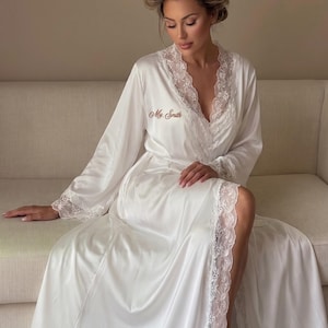 May include: A floor-length white satin robe featuring delicate white lace trim along the V-neckline and sleeve cuffs. The robe is embroidered with the text "Mrs. Smith" in a script font on the upper chest area, creating a refined and elegant look.
