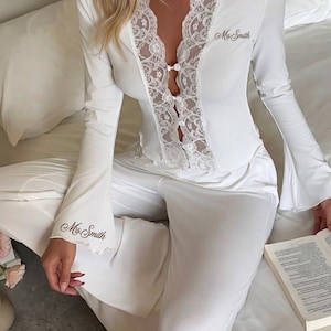 May include: A white long-sleeve pajama set featuring a button-down top with a lace-trimmed V-neckline and flared sleeves. The text "Mrs. Smith" is embroidered in dark thread on the left chest and the right sleeve cuff.