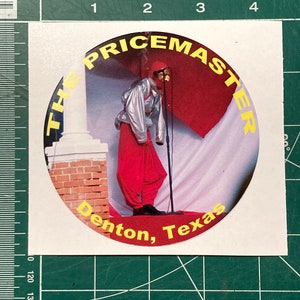 May include: A yellow and black circular sticker with the text "The Pricemaster" in a yellow circle. The sticker features a person wearing a red jumpsuit, a silver jacket, and a red hat, standing on a brick platform. The text "Denton, Texas" is printed in yellow at the bottom of the sticker.