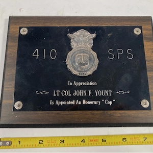 May include: A wooden plaque with a black center and silver accents. The plaque features the text "410 SPS" and "In Appreciation LT COL JOHN F. YOUNT Is Appointed An Honorary Cop". A silver badge is affixed to the plaque.