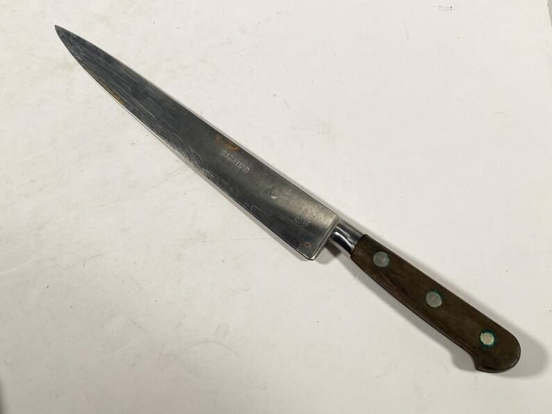 May include: A stainless steel carving knife with a dark brown wooden handle. The blade is long and slender, with a sharp point. The handle is attached with a metal bolster and secured with three silver rivets. The word 'SARADES' is etched on the blade.