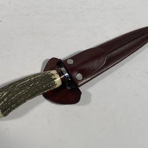 May include: A hunting knife with a textured antler handle and a brown leather sheath. The knife blade is silver, and the sheath is secured with two silver rivets. The handle has a cream-colored base and a dark green textured pattern.