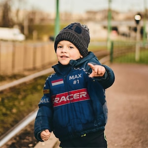 Può includere: Un bambino indossa un cappello di lana grigio scuro con un pompon e una giacca blu. La giacca ha la scritta "RACER" e il nome "MAX #1" stampati, insieme a strisce rosse e bianche e un disegno della bandiera olandese.