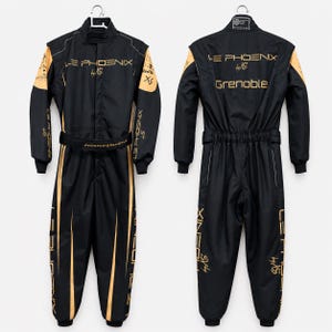 May include: A black racing jumpsuit with gold accents, featuring the text LE PHOENIX by XS and GRENOBLE. The garment includes a high collar, long sleeves, a belted waist, and full-length legs, shown from both the front and back views on hangers.