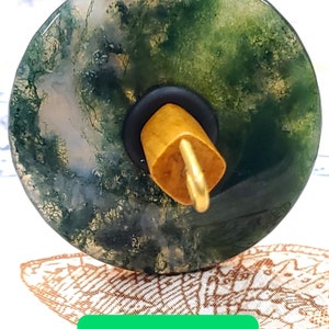 May include: A spinning wheel whorl made of moss agate with a green, brown, and white pattern. The whorl is attached to a wooden spindle with a gold metal hook.