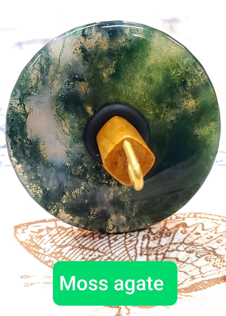 May include: A spinning wheel whorl made of moss agate with a green, brown, and white pattern. The whorl is attached to a wooden spindle with a gold metal hook.