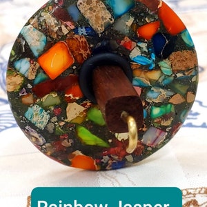 May include: A spinning wheel whorl with a rainbow jasper design. The whorl is made of a dark green base with colorful flecks of jasper in shades of orange, red, blue, green, and brown. The whorl is attached to a wooden spindle with a black rubber ring and a gold hook.