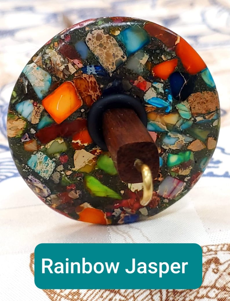 May include: A spinning wheel whorl with a rainbow jasper design. The whorl is made of a dark green base with colorful flecks of jasper in shades of orange, red, blue, green, and brown. The whorl is attached to a wooden spindle with a black rubber ring and a gold hook.
