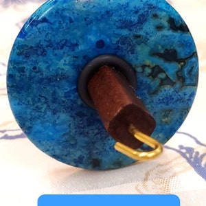 May include: A spinning wheel with a blue lace agate whorl, a dark brown wooden spindle, and a gold metal hook.