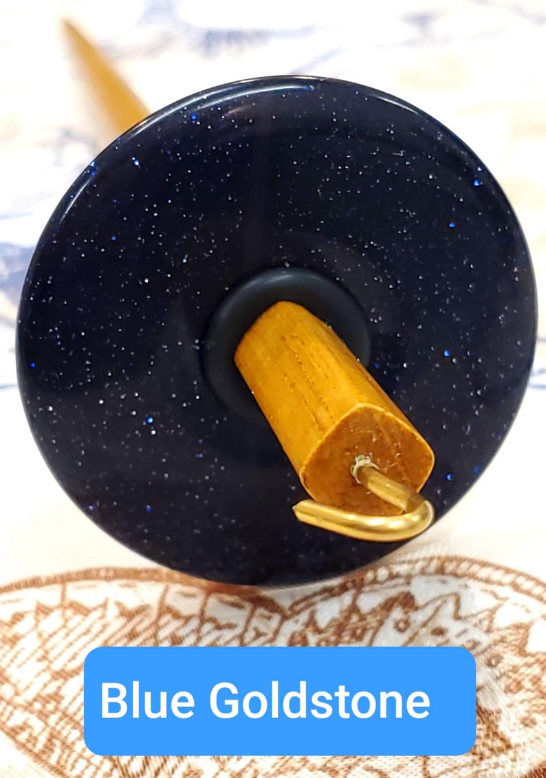 May include: A spinning wheel with a blue goldstone whorl and a wooden spindle. The whorl has a sparkly, glittery finish.