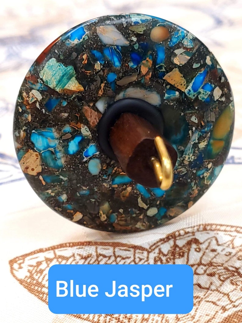 May include: A spinning wheel whorl made of blue jasper with a brown wooden spindle and a gold metal hook. The whorl has a speckled pattern of blue, brown, and white.