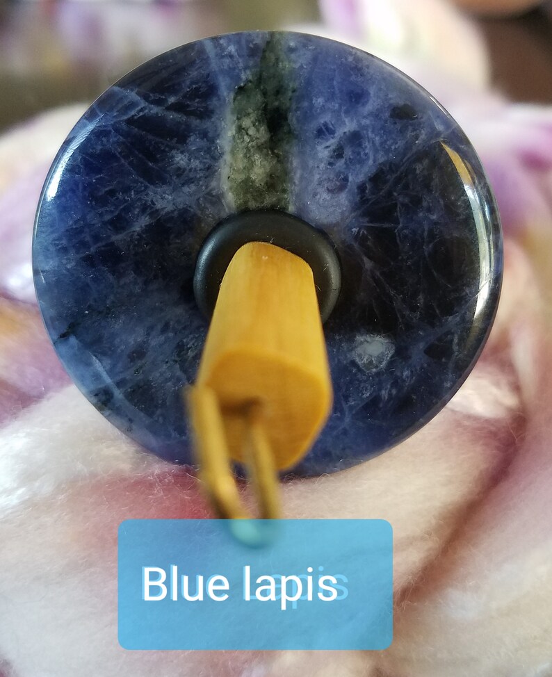 May include: A spinning wheel whorl made of blue lapis lazuli stone. The whorl is round and has a dark blue color with white and black veins. The whorl is attached to a wooden spindle.