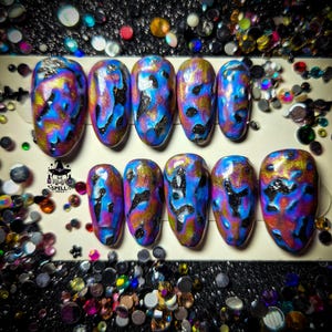 Può includere: Un set di dieci unghie finte con un design astratto vibrante. Le unghie presentano una base di tonalità blu e viola, accentuata da motivi oro e neri. L'immagine include il testo "SPELL NAILS".